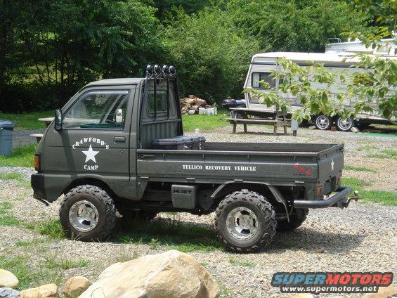 Olive drab mini truck with 'CRAWFORD'S CAMP' and 'TELLICO RECOVERY VEHICLE' text, off-road tires, and a camper trailer in background