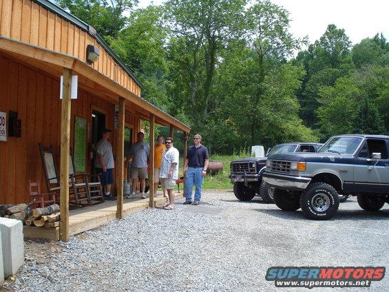 Two lifted Ford F-Series trucks parked outside a wooden building with people on the porch