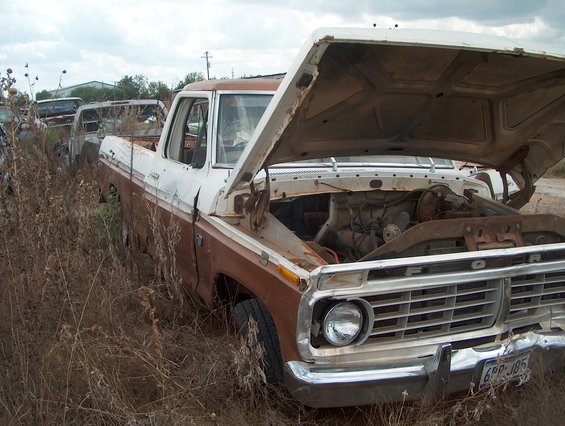 Old Ford pickup truck with hood open, parked in a junkyard