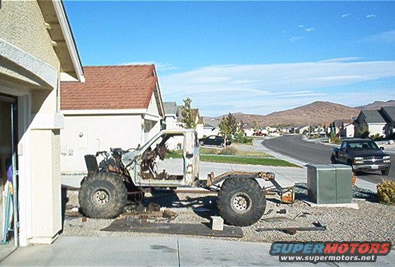 Modified truck chassis with large tires, side view. Another truck drives by