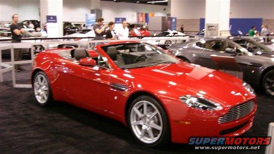 Red convertible sports car with silver wheels, parked indoors. Another dark car is visible behind it