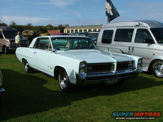 White 1965 Pontiac Catalina coupe parked on grass, front 3/4 view, with a van and other vehicles in the background