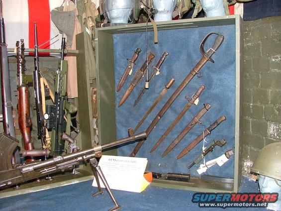 Display of firearms and bayonets, including a machine gun and swords, with a helmet on the right
