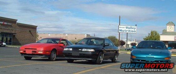 Red, black, and teal coupes parked in a lot. The black one looks like a 1989 Mazda MX-6