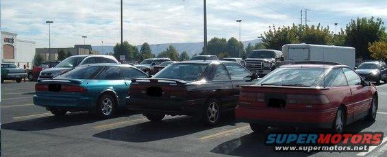 Three coupes parked in a lot: teal, black, and red. The red car is a 1989 Ford Probe