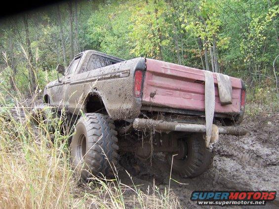 Red pickup truck with large rear tires, seen from behind on a dirt road