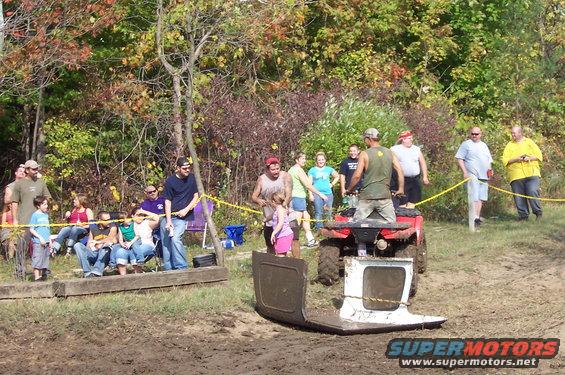 People watch an ATV pull a car door through mud; trees and spectators in background