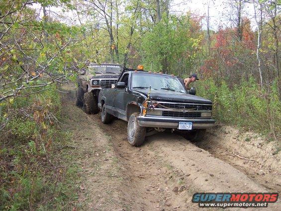 Black pickup truck and an older SUV on a muddy trail, with a person leaning into the truck