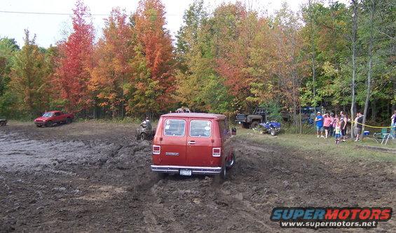 Red van driving through mud pit with fall foliage in background