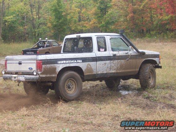 Muddy white pickup truck with black stripe and snorkel, driving off-road. Another Toyota pickup is visible behind it