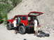 1996 Jeep Cherokee - Two people load gear into a red Jeep Cherokee parked on a dirt road with a rocky hillside in the background.