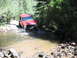 1996 Jeep Cherokee - A red Jeep Cherokee navigates a rocky stream crossing on a dirt path surrounded by dense foliage.