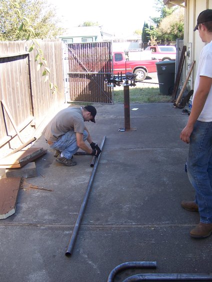 Two people working on metal pipes in a backyard with a red pickup truck and a Bronco in the background