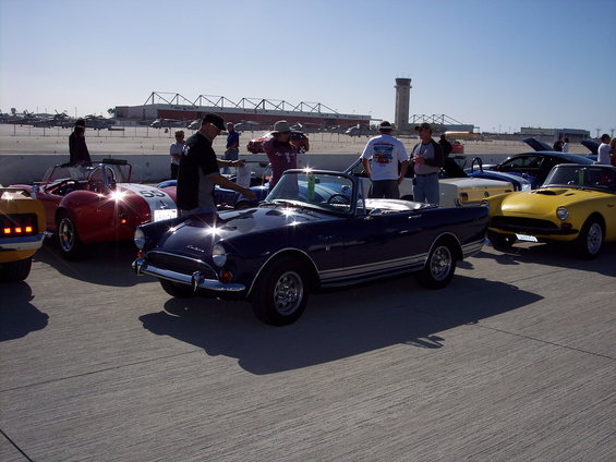 Dark blue convertible sports car with silver side stripes, parked outdoors with other classic cars