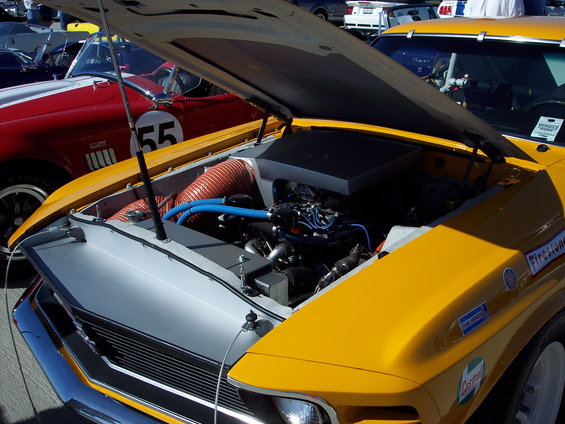 Engine bay of a yellow muscle car with hood open, showing V8 engine and orange intake hose. Red car with number 55 visible in background