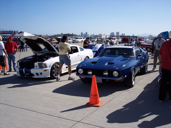Blue muscle car with hood scoops and racing stripes, parked next to a white modern Mustang with its hood open