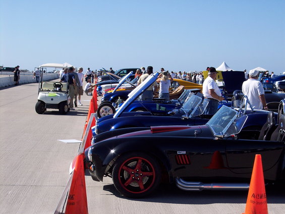 Several classic sports cars are parked in a line on a paved surface under a clear blue sky. A golf cart is visible to the left
