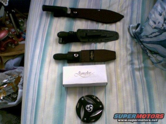 Three knives in sheaths and a white box with text are laid out on a striped blanket
