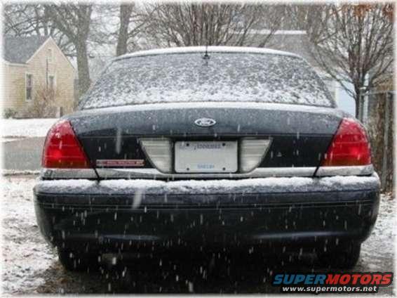 Rear view of a dark Ford Crown Victoria covered in snow, with a house and bare trees in the background