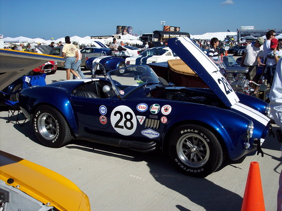 Blue race car with white stripes, number 28, and various sponsor decals. Hood is open, showing engine details. Other cars and tents are visible in the background