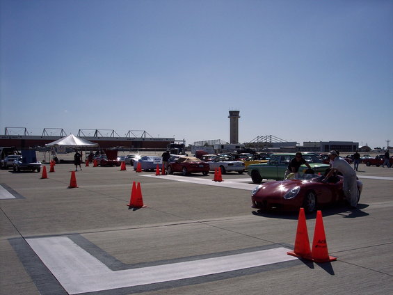red convertible sports car with two people near the driver's side, parked on an airfield with other cars and a tent in the background