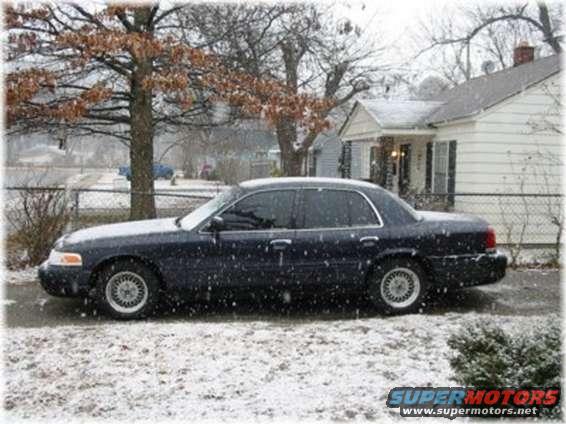 Dark sedan parked on a snowy road, side view. The car has tinted windows and silver wheels