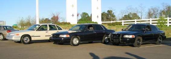 Three Ford Crown Victoria police cars parked side-by-side. The leftmost car is tan, the other two are black