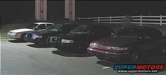 Four cars parked in a lot at night: a white sedan, two dark sedans with push bars, and a maroon sedan