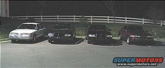 four sedans parked in a lot at night, one white and three dark colored
