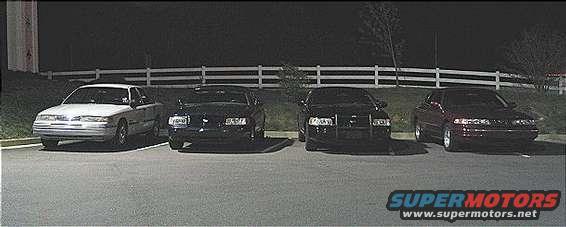 Four cars parked in a lot at night. The first car is white, the other three are dark. The 1999 Ford Crown Victoria is visible
