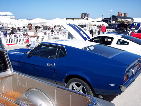 Blue coupe with white roof and racing stripes on hood, parked next to white car with stripes. Outdoor event with tents and people