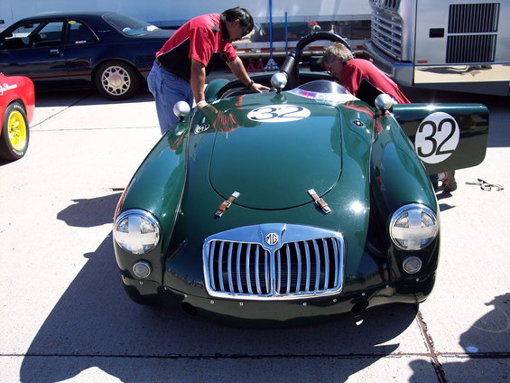 Green MG race car with number 32 on hood and door, two men looking inside. Dark car and red car in background