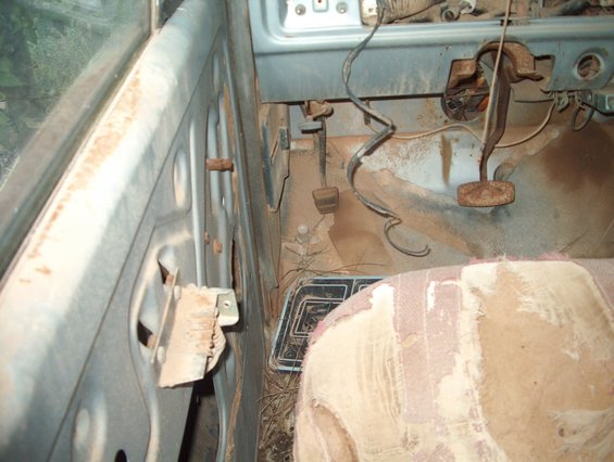 Interior of a rusty, dusty 1978 Ford Bronco showing the driver's side door panel, pedals, and torn seat