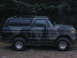1989 Ford Bronco - A camouflaged Ford Bronco is parked on a dirt road with trees in the background.