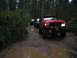 1989 Ford Bronco - Several off-road vehicles navigate a wet and muddy path through a dense forest.