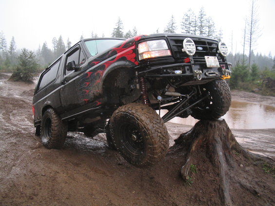 1989 Ford Bronco with flame decals driving on a muddy hill, front tire on a tree stump
