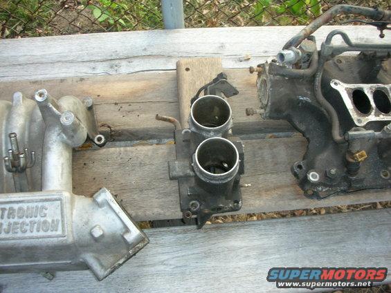 Engine intake manifold and throttle body parts on a wooden surface. Text reads 'ELECTRONIC INJECTION'
