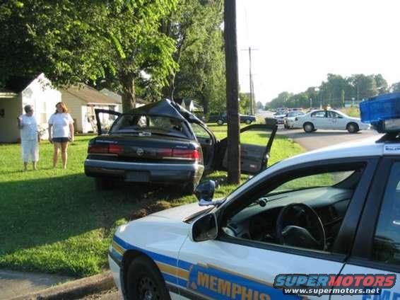 wrecked black sedan with doors open, leaning against a pole, police car in foreground, two women standing nearby