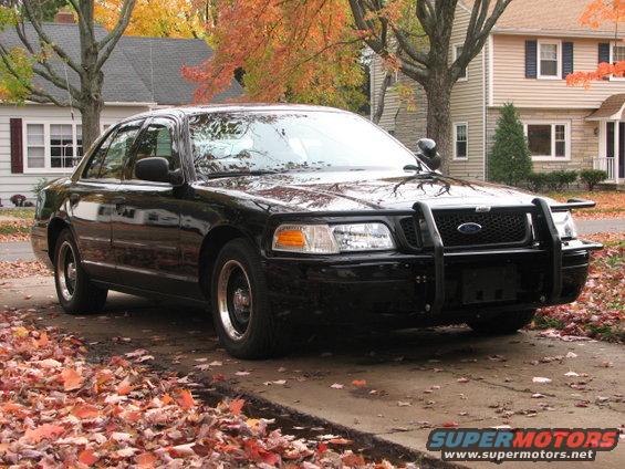 Black 2000 Ford Crown Victoria police car with a push bar, parked on a driveway with fallen leaves