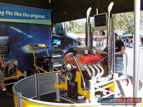 Chevrolet 572 engine on display with chrome exhaust pipes and a screen above