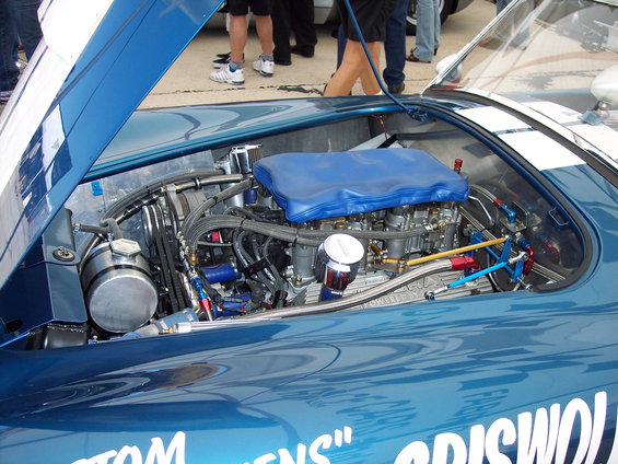 Blue race car engine bay with V8 engine, dual carburetors, and custom plumbing. Text on fender reads "TOM GENS" GRISWOLD"