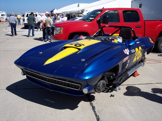 Blue race car with yellow stripe and number 26, missing wheels, on jacks. Red truck and people in background