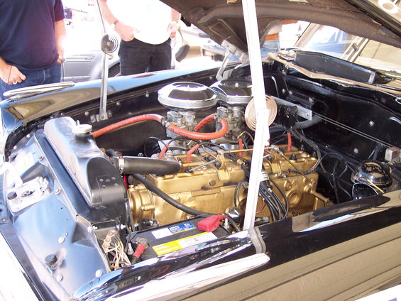 Engine bay of a classic car showing a gold-colored V8 engine with dual carburetors and red hoses