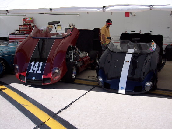 Two race cars, one red with number 141, the other black with a white stripe, parked under a tent