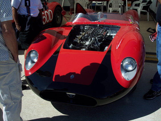 Red and black race car, front view, engine exposed, with a person to the left and another car in the background