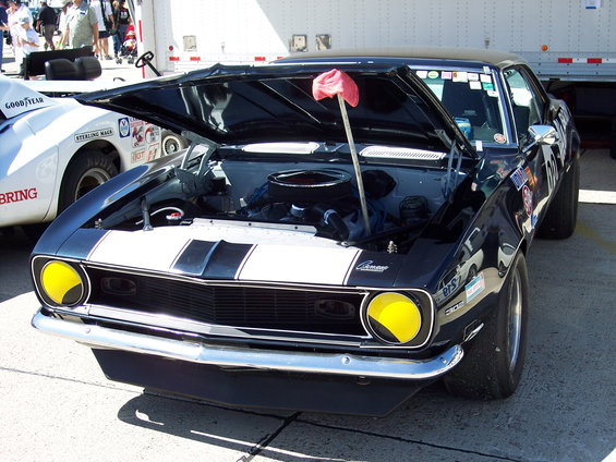 Black 1969 Chevrolet Camaro SS with white racing stripes and yellow headlights, hood open. Another white car is partially visible to the left