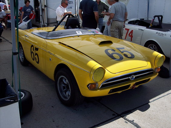 Yellow convertible race car with number 65 on hood and door, parked next to a white car with number 74