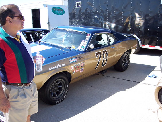 Gold and blue race car with number 78, Goodyear tires, and various sponsor decals. Man in striped shirt stands nearby. Trailer in background