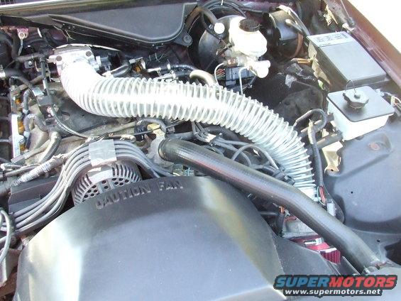Engine bay showing a large flexible tube cold air intake system installed. Fan shroud has 'CAUTION FAN' text