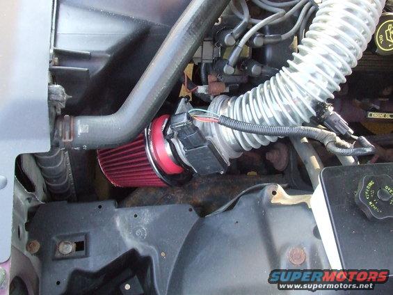 Engine bay showing a red cone air filter connected to a ribbed intake hose. Some wiring and hoses are visible